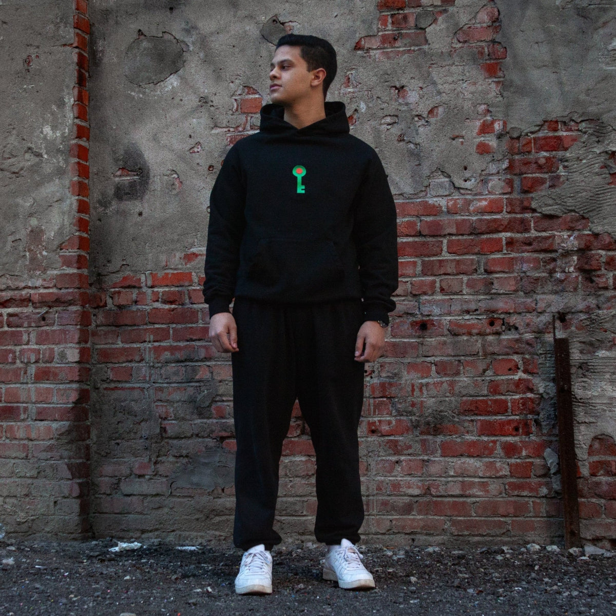 Model wearing the black ‘Key of Return’ hoodie, a design inspired by Palestinian heritage, featuring an embroidered green key symbolizing the right of return, standing against a textured brick wall.