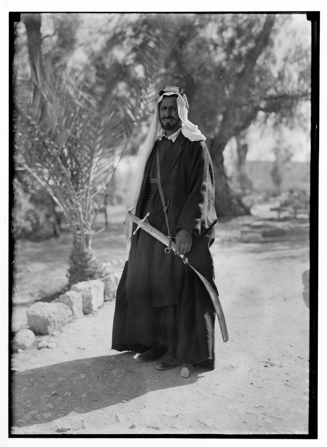 A Bedouin sheikh in Palestine standing in a dark wool cloak worn open over his robe, with a keffiyeh and agal on his head