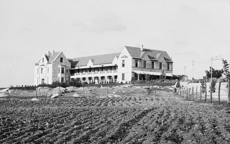 The Kadoorie Agricultural School at Tulkarm in the early 1930s, a long stone building with an arcaded veranda standing above ploughed furrows and a wire fence