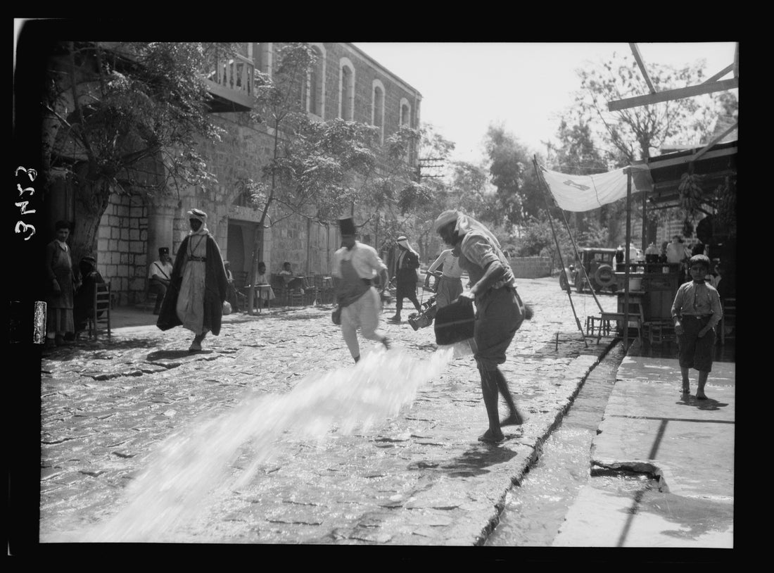 The cobbled main street of Beisan around 1920, with stone shopfronts, market awnings and townspeople, as water is sluiced across the paving from an open gutter