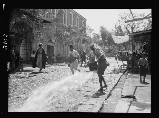 The cobbled main street of Beisan around 1920, with stone shopfronts, market awnings and townspeople, as water is sluiced across the paving from an open gutter