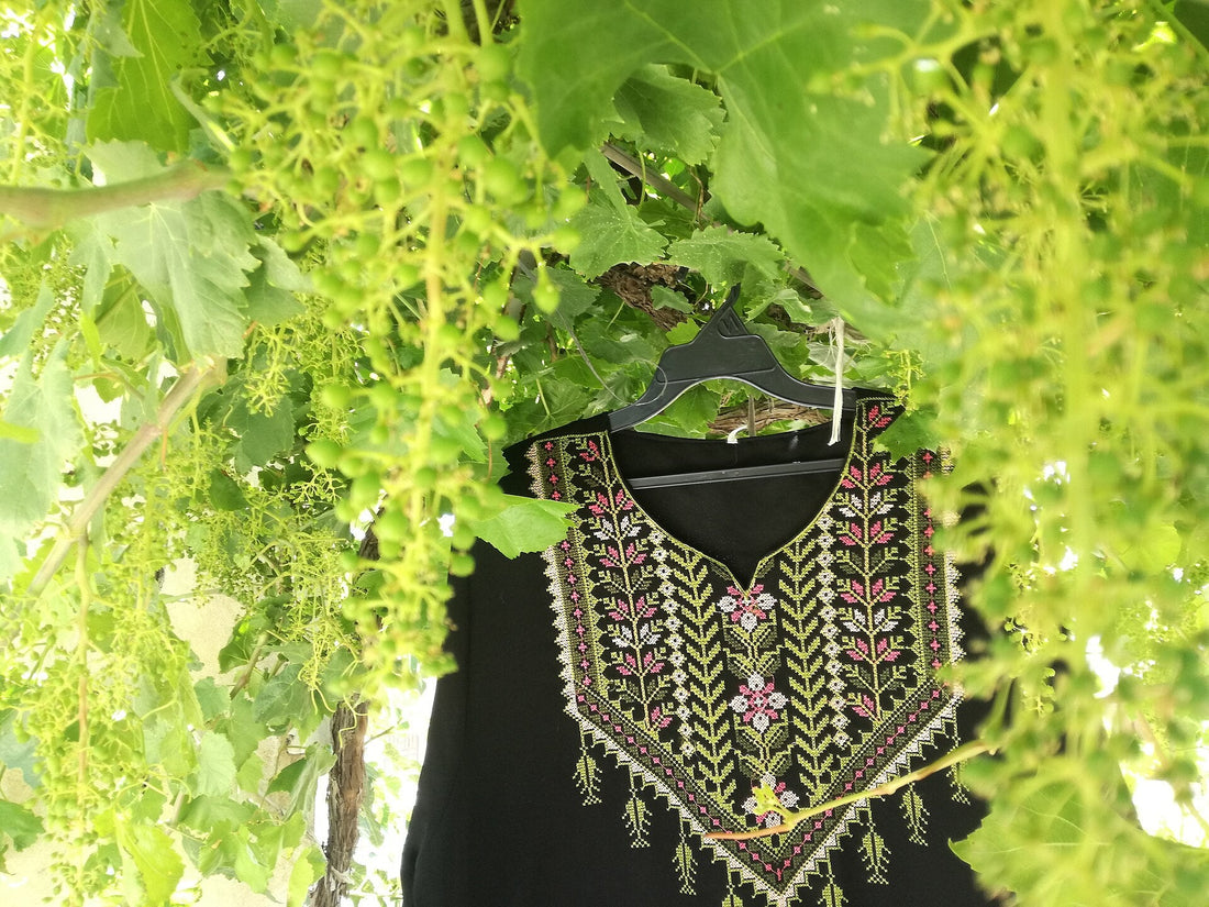 A Palestinian thobe's embroidered qabbeh chest panel in pink, white and olive cross-stitch, hanging beneath grapevine leaves