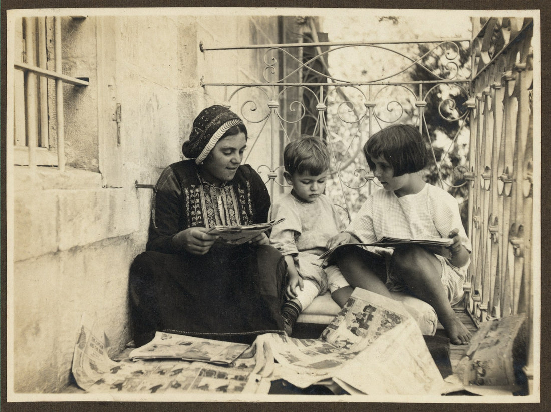 A woman in embroidered Palestinian dress sits with two young children on a stone balcony, all three looking at illustrated papers together