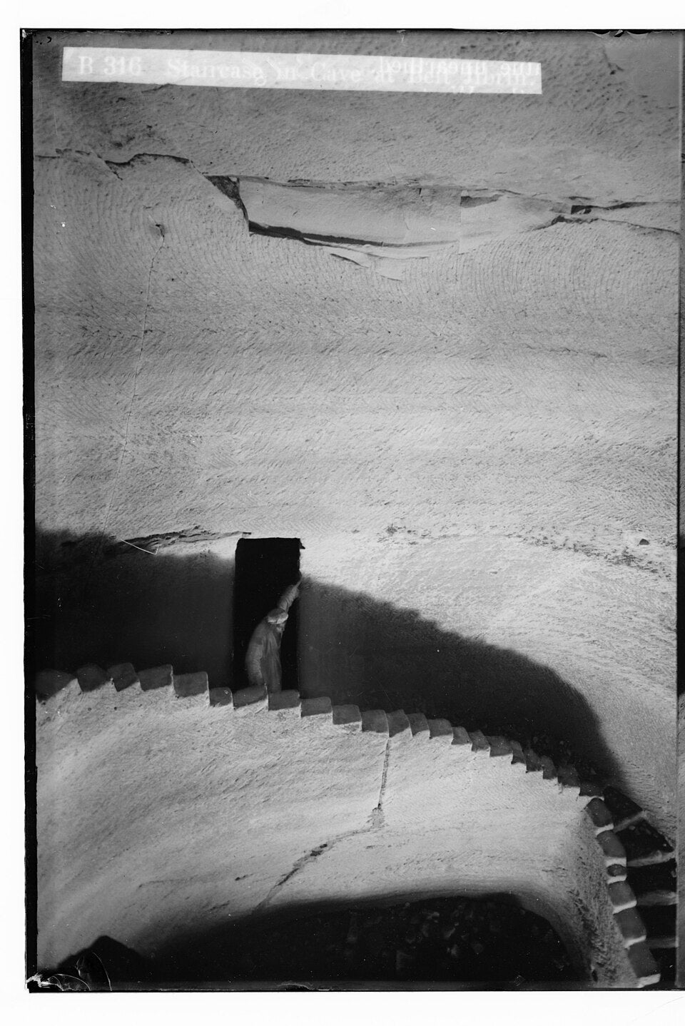 A carved stone spiral staircase descending into a bell-shaped cave at Beit Jibrin, Palestine, with a figure on the stairs for scale