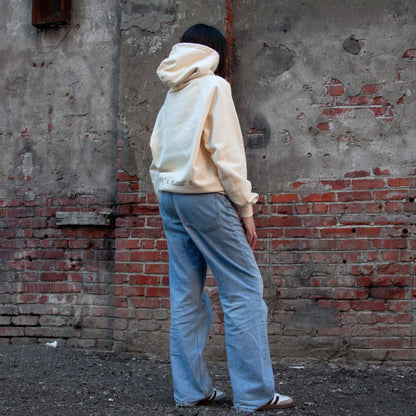 model wearing a sand-colored hoodie with coordinates symbolizing Palestine embroidered on the back, standing in front of a rustic brick wall.