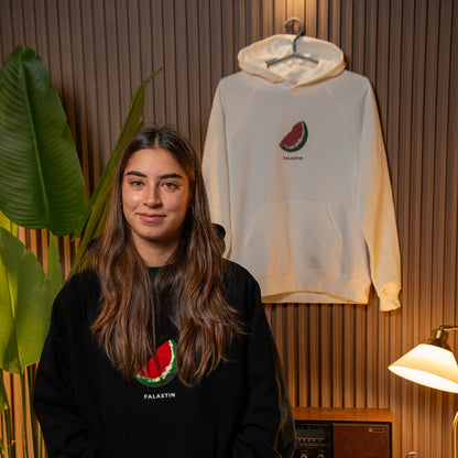 Person wearing a black hoodie with a watermelon design, standing next to a white hoodie with a similar design on a wooden wall.