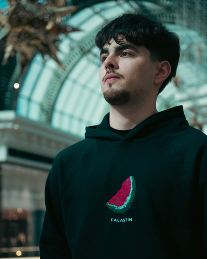 Person wearing a dark hoodie with a watermelon design and 'FALASTIN' text, standing in a decorative indoor setting.