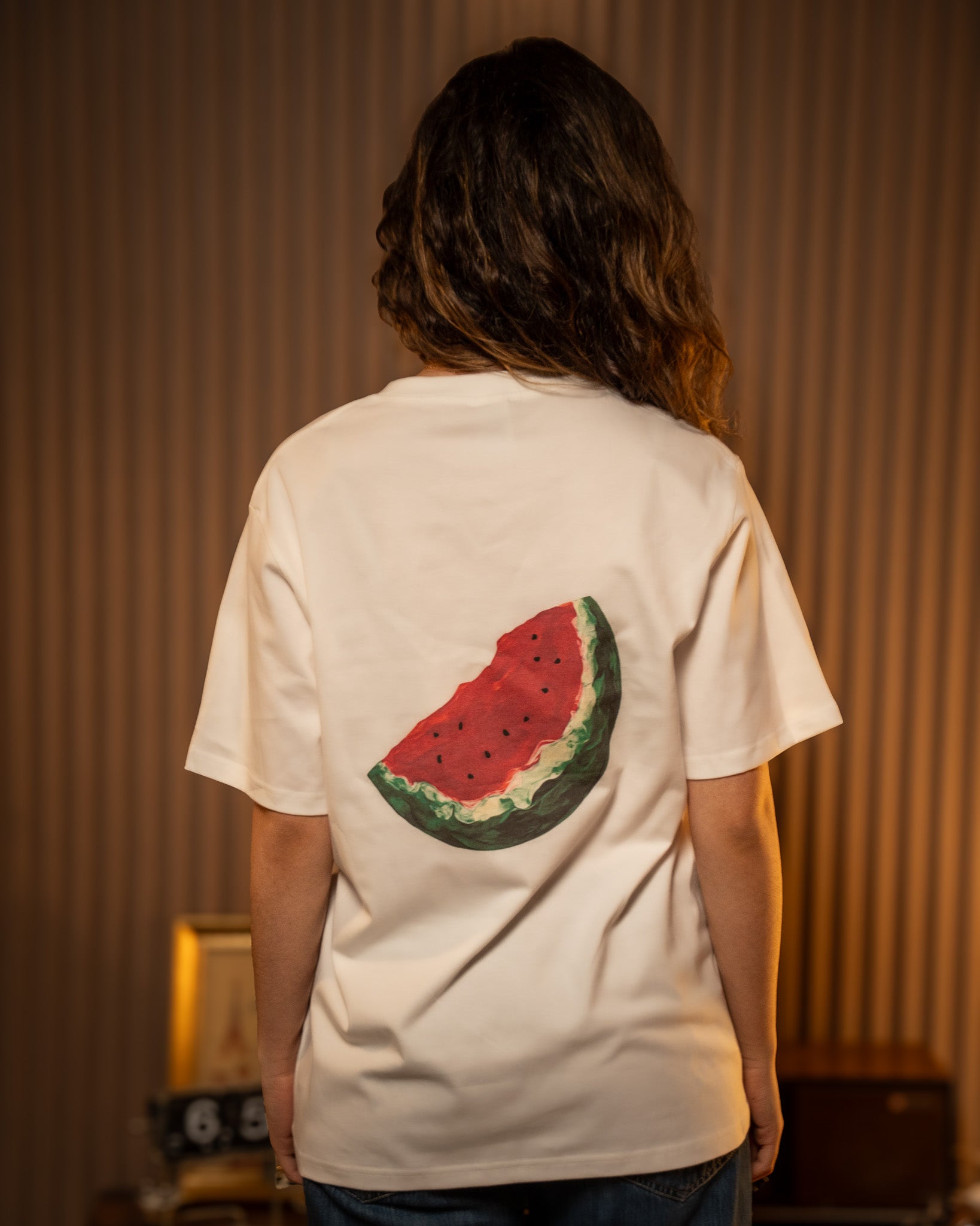 Person wearing a white t-shirt with a Palestinian watermelon design on the back, standing against a neutral background.