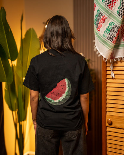 Person wearing a black t-shirt with a Palestinian watermelon design on the back, standing in a room with plants and a wooden cabinet.