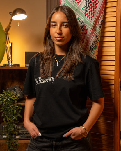 Woman wearing a Palestinian black t-shirt with text explaining the symbol of the Palestinian watermelon, standing in a room with decorative elements.