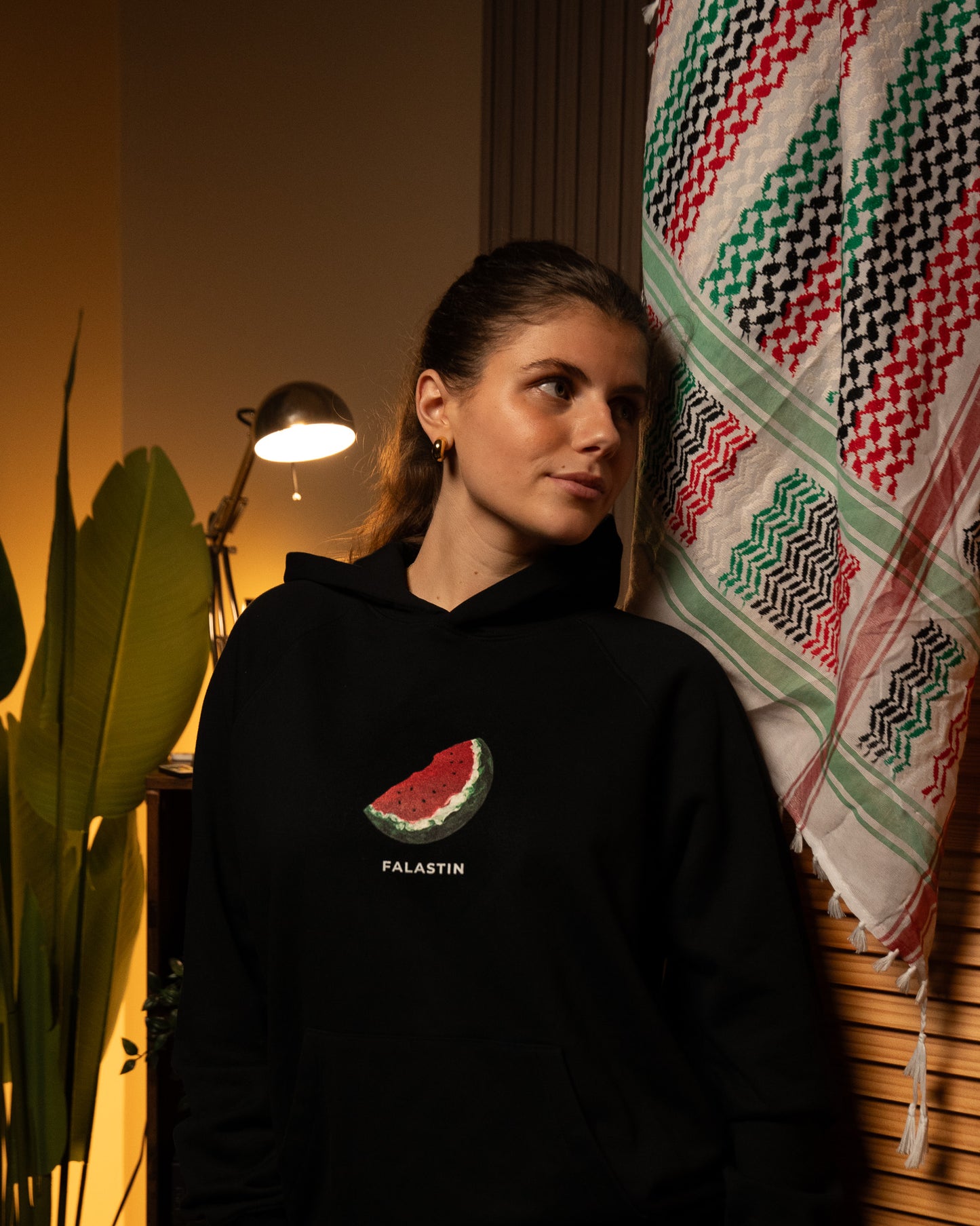 Person wearing a black hoodie with a Palestinian watermelon design and 'FALASTIN' text, standing in a room with a lamp and patterned towel.