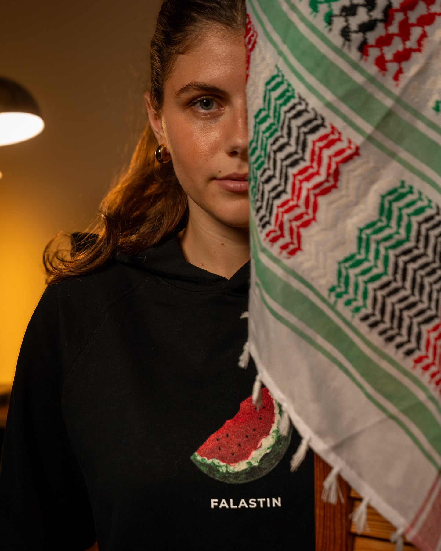 Person wearing a black hoodie with a Palestinian watermelon design and 'FALASTIN' text, standing next to a colorful patterned towel.