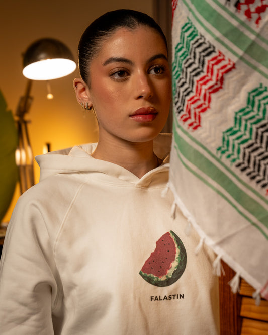 Person wearing a white hoodie with a Palestinian watermelon design and 'FALASTIN' text, holding a colorful Palestinian keffiyeh.