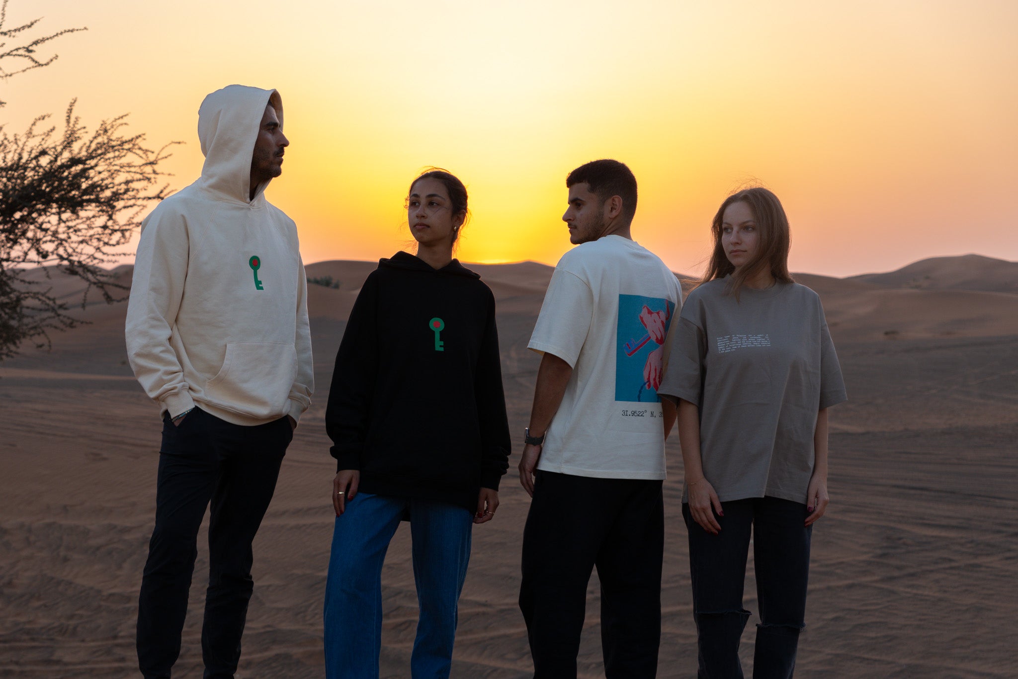 Four models standing in a desert at sunset wearing FALASTIN apparel, including the Key of Return hoodies in cream and black, and the Key of Return t-shirts in sand and white.