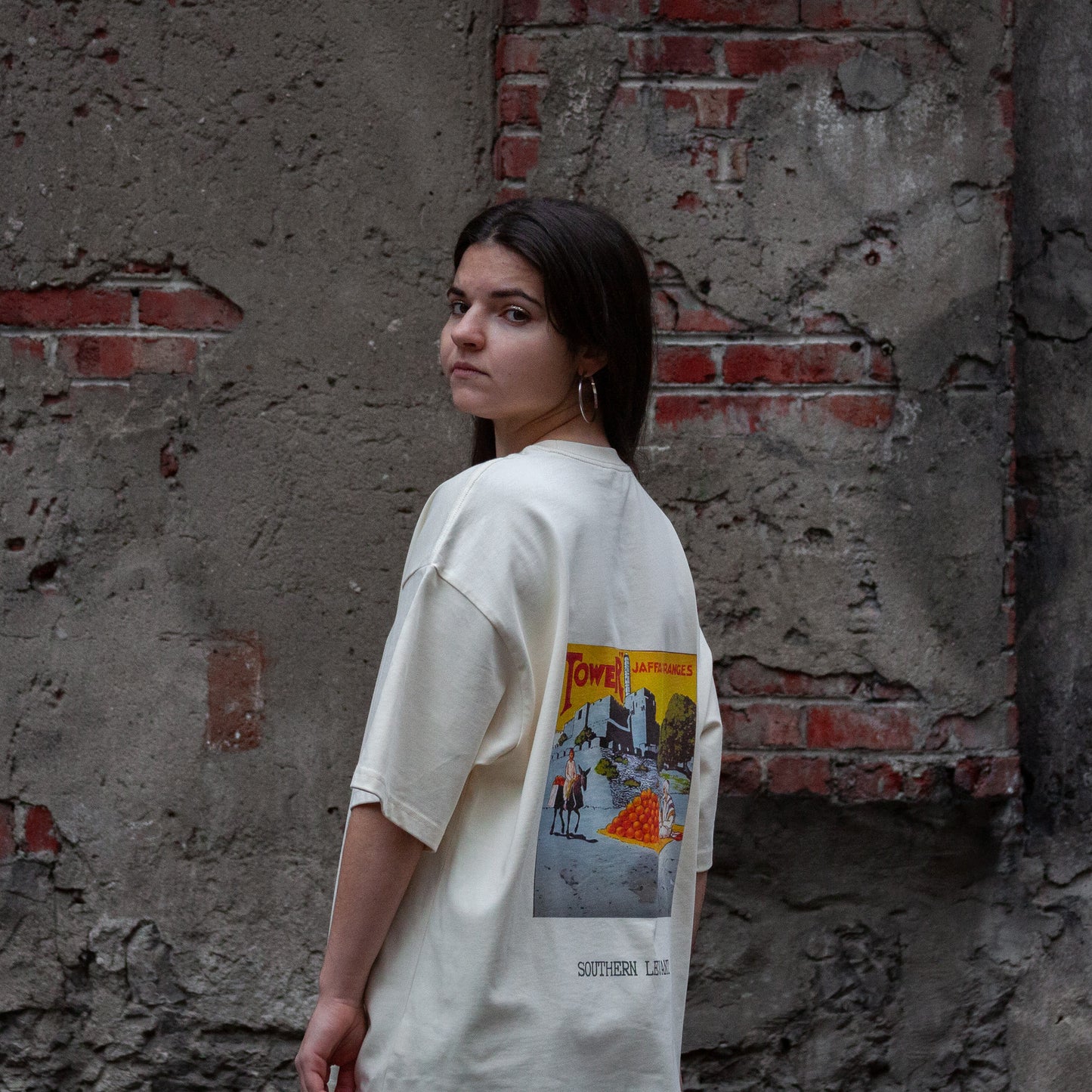 A model wearing the Jaffa Orange T-Shirt in Sand from FALASTIN, showcasing the back design featuring a vintage-inspired graphic of Jaffa oranges and the text “Southern Levant.” Set against a rustic brick wall background.