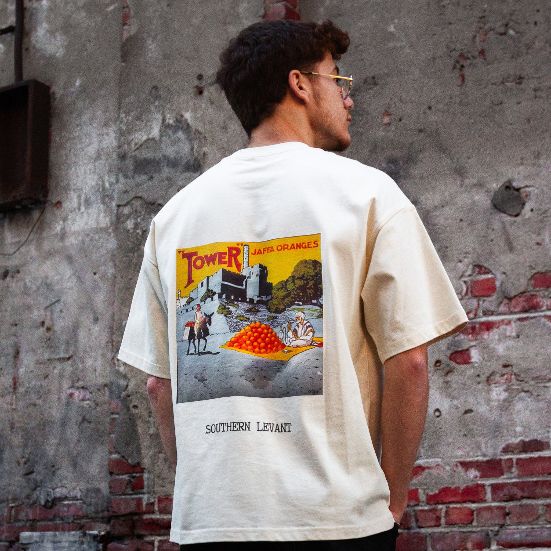 A model wearing the Jaffa Orange T-Shirt in Sand from FALASTIN, showcasing the back design featuring a vintage-inspired graphic of Jaffa oranges and the text “Southern Levant.” Set against a rustic brick wall background.