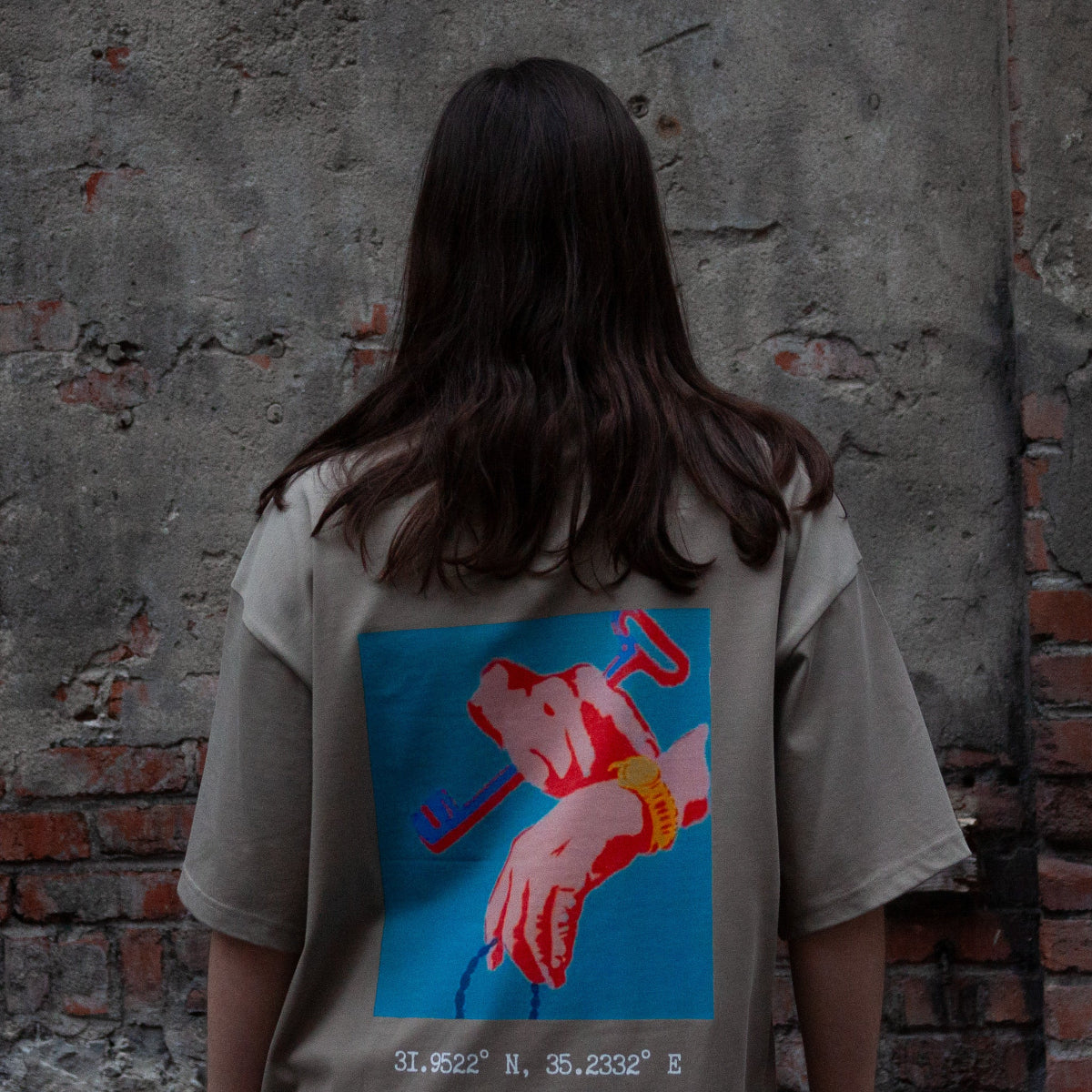 Back view of a model wearing the FALASTIN Key of Return T-Shirt in coffee color, featuring a bold graphic design of hands holding a key and the geographical coordinates of Palestine.