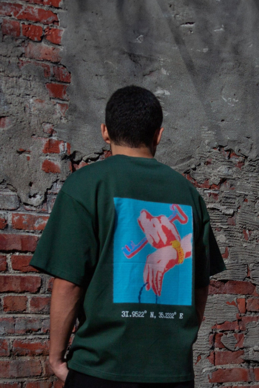 Back view of a model wearing the FALASTIN Key of Return T-Shirt in forest color, featuring a bold graphic design of hands holding a key and the geographical coordinates of Palestine.