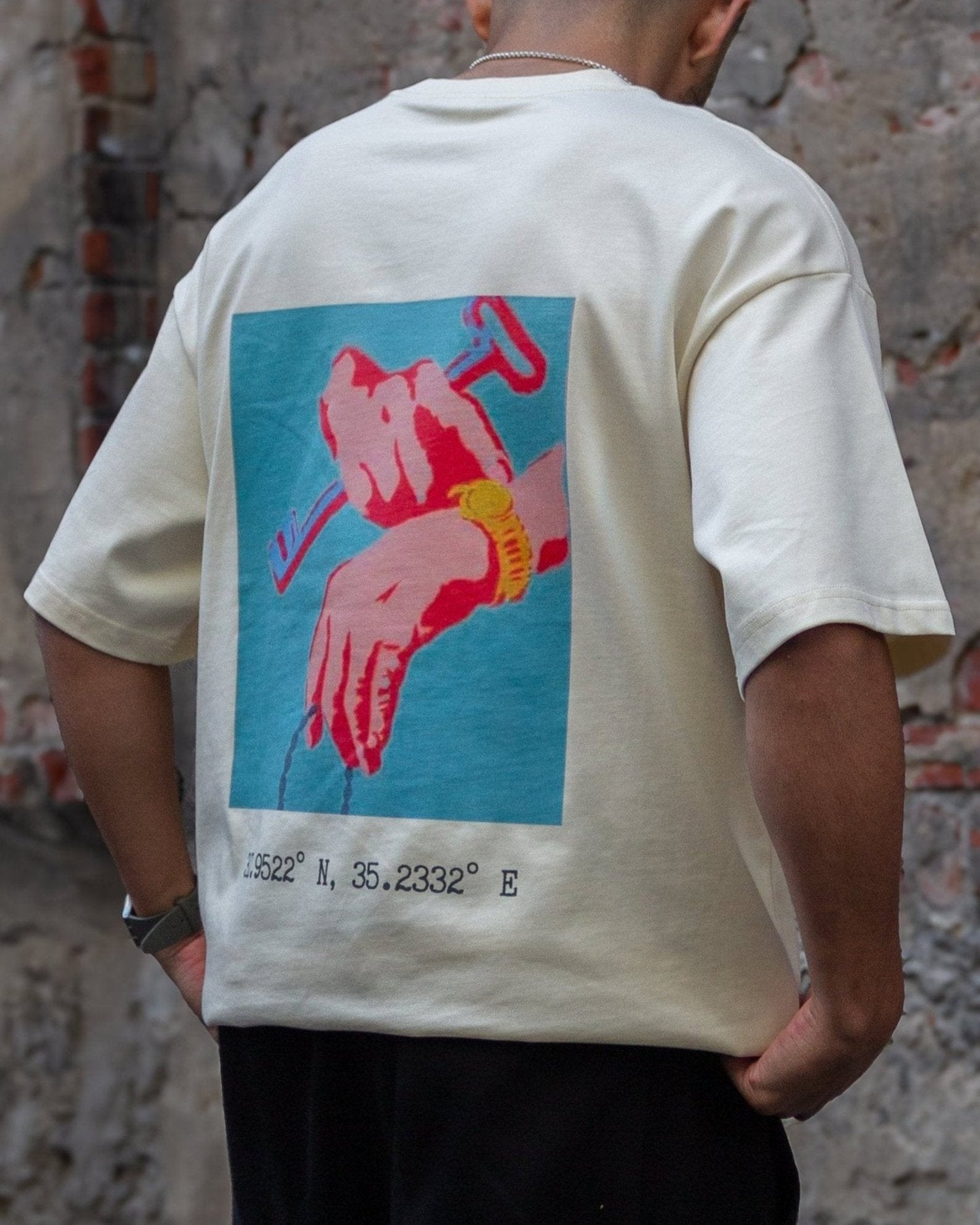 Back view of a model wearing the FALASTIN Key of Return T-Shirt in sand color, featuring a bold graphic design of hands holding a key and the geographical coordinates of Palestine.