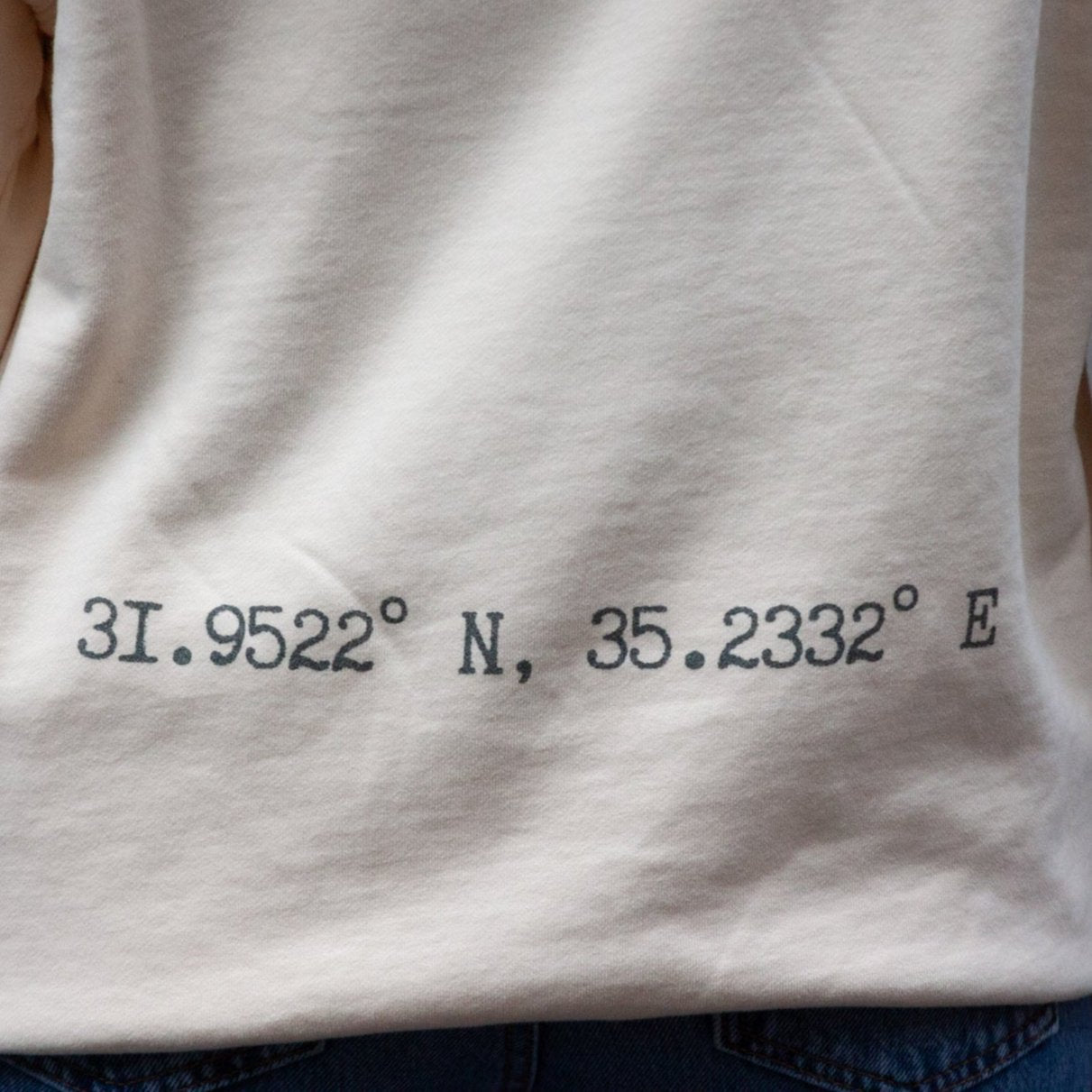 Close-up of embroidered geographic coordinates, 31.9522° N, 35.2332° E, symbolizing the location of Palestine on a sand-colored hoodie.