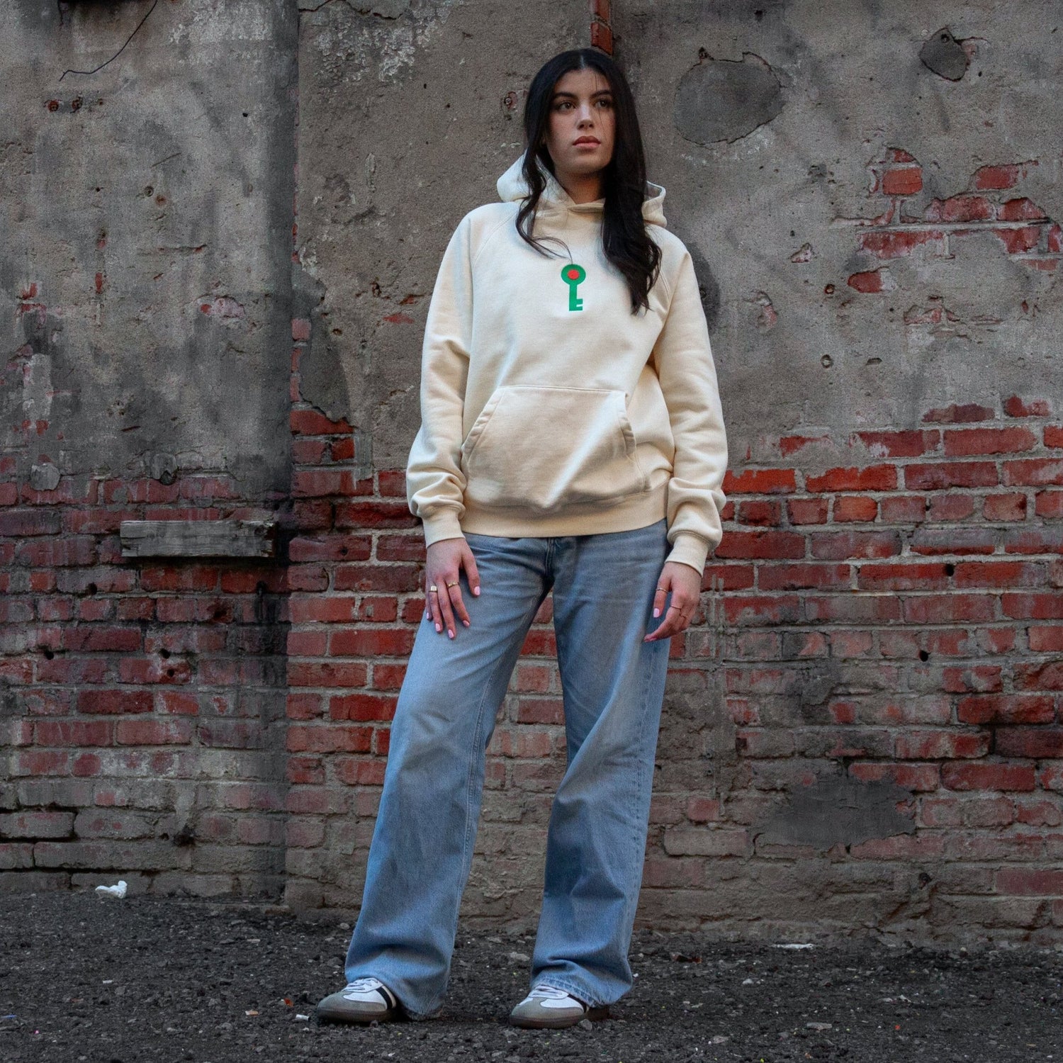 Front view of model wearing a sand-colored hoodie featuring an embroidered green key with a red circle emblem , symbolizing Palestinian heritage, standing in front of a rustic brick wall.