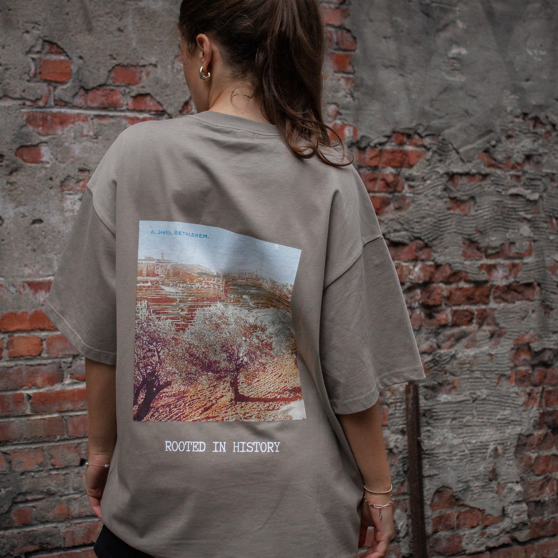 A model wearing the Heritage Olive Tree T-Shirt in Coffee from FALASTIN, showcasing the back design featuring an olive tree graphic and the words “Rooted in History.” Set against a rustic brick wall background.