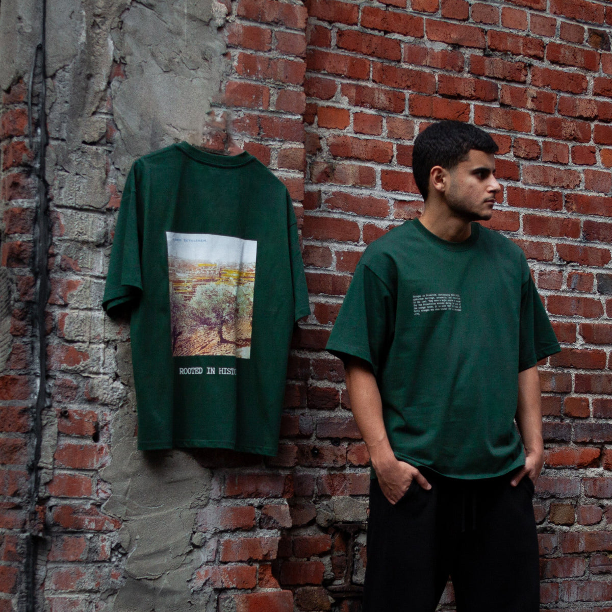 Model wearing the Heritage Olive Tree T-Shirt in forest green, featuring text on the front that highlights the olive tree’s deep connection to Palestinian heritage. The same shirt is hung on a red brick wall, showcasing the back design: a graphic of an olive grove from Bethlehem with the words ‘Rooted in History’ beneath.