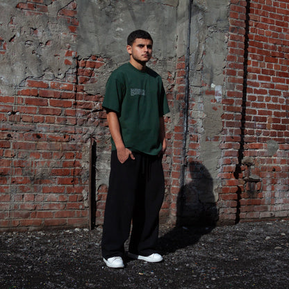 A model wearing the Heritage Olive Tree T-Shirt in Forest from FALASTIN, showcasing the front design featuring small text explaining the significance of the Palestinian olive tree symbol. Set against a rustic brick wall background.
