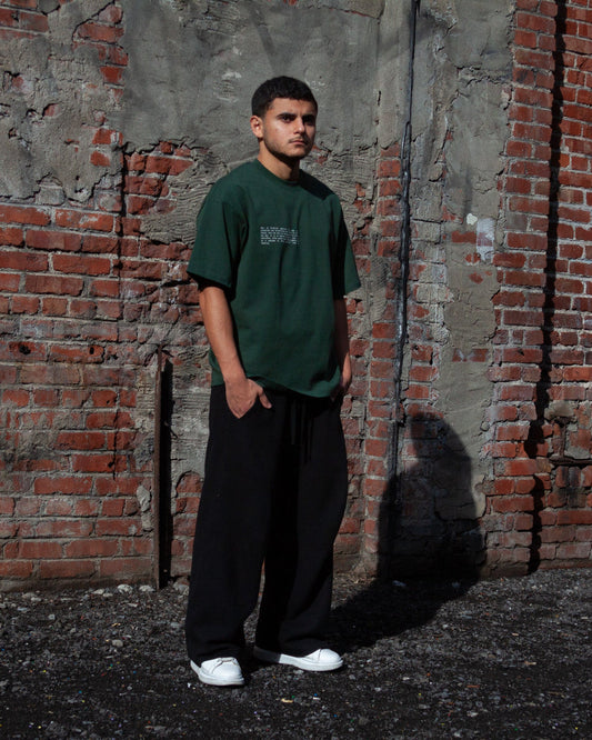 A model wearing the Heritage Olive Tree T-Shirt in Forest from FALASTIN, showcasing the front design featuring small text explaining the significance of the Palestinian olive tree symbol. Set against a rustic brick wall background.