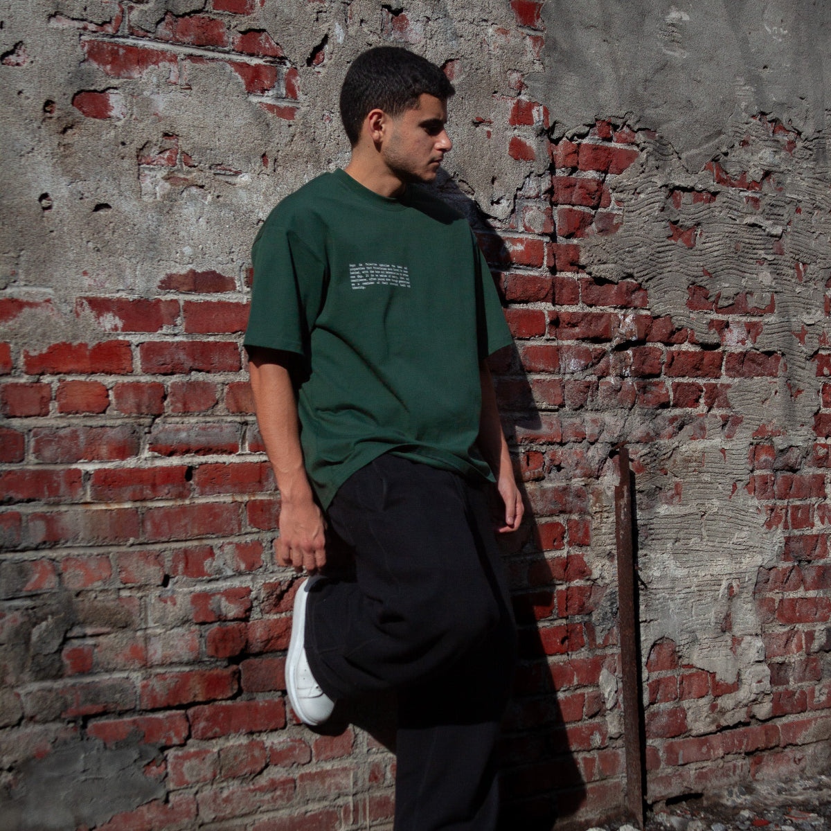 A model wearing the Heritage Olive Tree T-Shirt in Forest from FALASTIN, showcasing the front design featuring small text explaining the significance of the Palestinian olive tree symbol. Set against a rustic brick wall background.