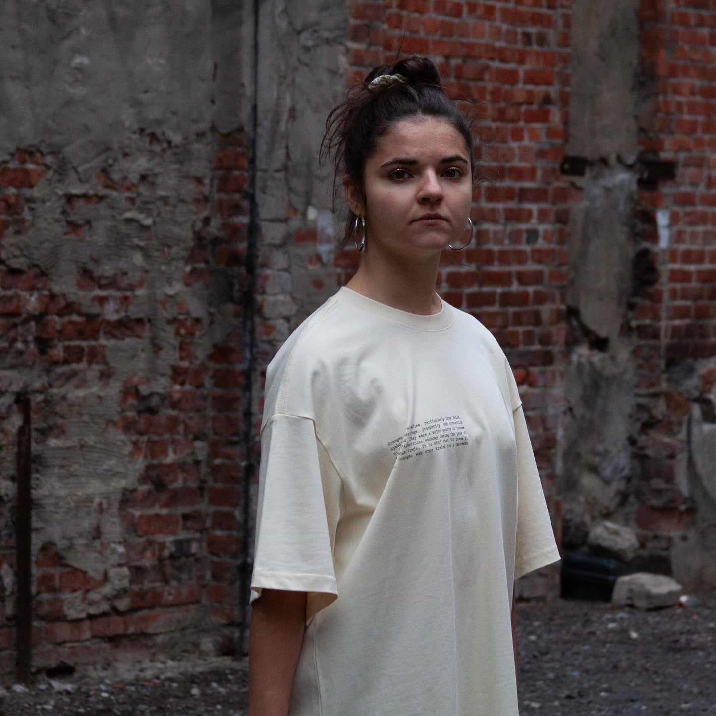 A model wearing the Heritage Olive Tree T-Shirt in Coffee from FALASTIN, showcasing the front design featuring small text explaining the significance of the Palestinian olive tree symbol. Set against a rustic brick wall background.