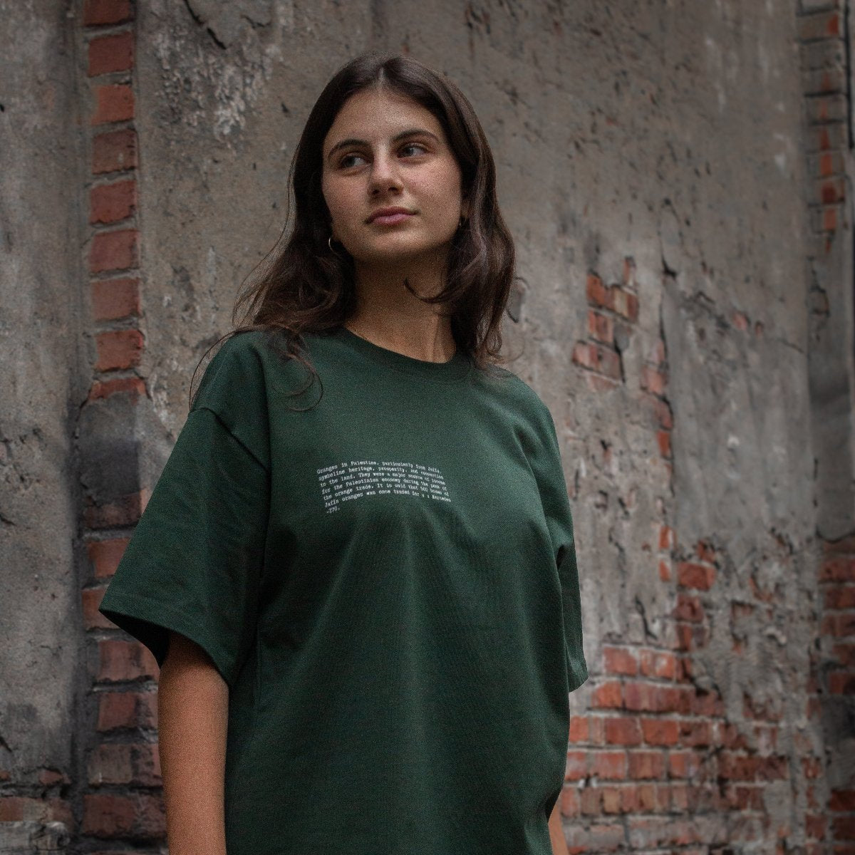 A model wearing the Jaffa Orange T-Shirt in forest from FALASTIN, featuring front text that highlights the cultural and historical significance of Palestinian oranges. The rich forest green color contrasts beautifully with the rustic brick wall backdrop, emphasizing the deep connection to Palestinian heritage.