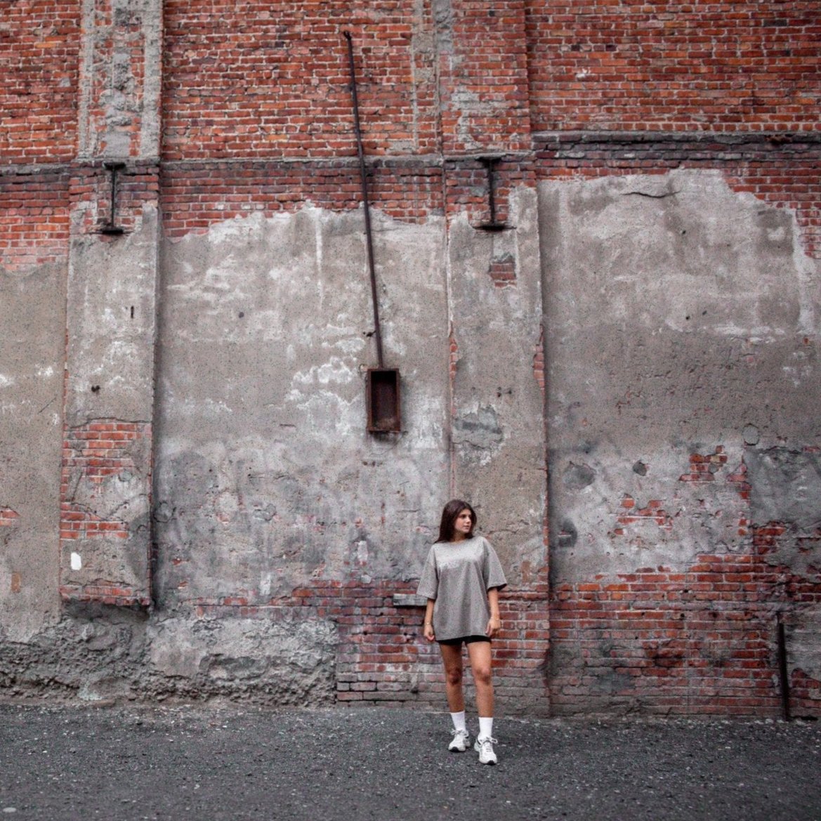 Model wearing the Heritage Key T-Shirt in coffee, shown in a far shot front view against a weathered red brick wall. The front of the shirt features text that emphasizes the cultural significance of the key symbol in Palestinian heritage.