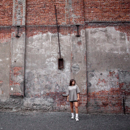 Model wearing the Heritage Key T-Shirt in coffee, shown in a far shot front view against a weathered red brick wall. The front of the shirt features text that emphasizes the cultural significance of the key symbol in Palestinian heritage.