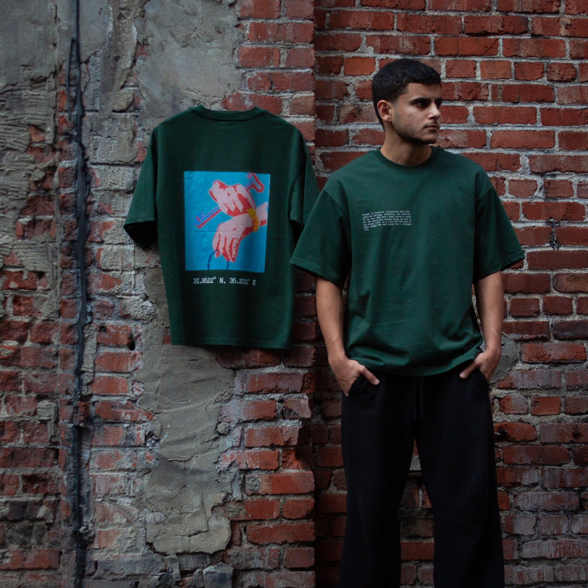 Model wearing the Heritage Key T-Shirt in forest green, featuring text on the front that explains the cultural significance of the key symbol in Palestinian heritage. The same shirt is hung on a red brick wall, showcasing the back design: a historic graphic of a key with geographic coordinates printed underneath